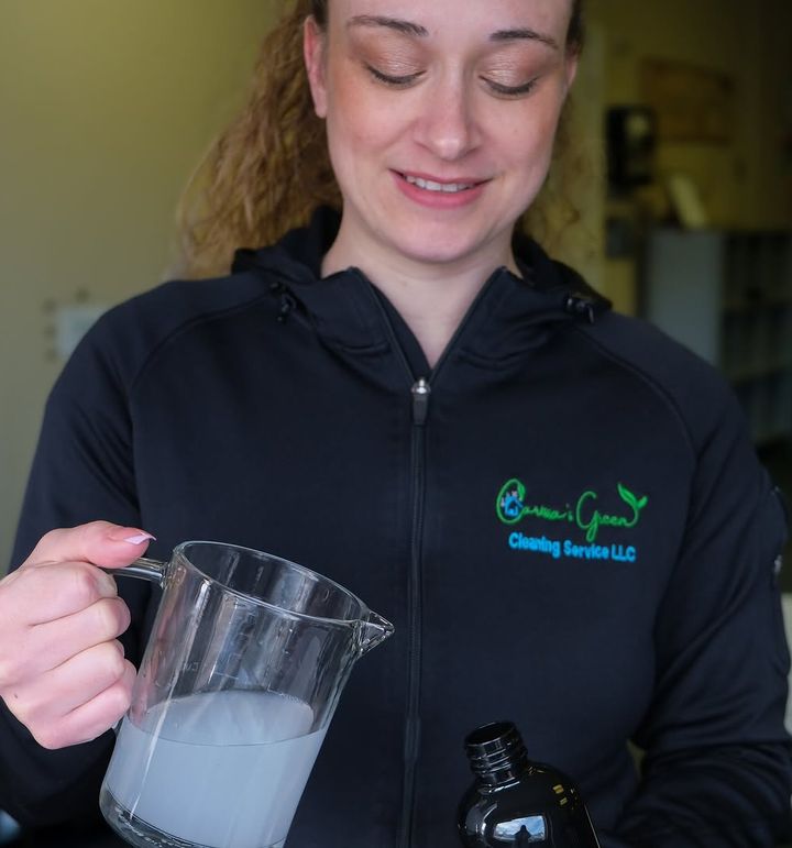 A woman in a black zip jacket with the Carissa's Green logo pouring a cloudy cleaning solution from a measuring jug.